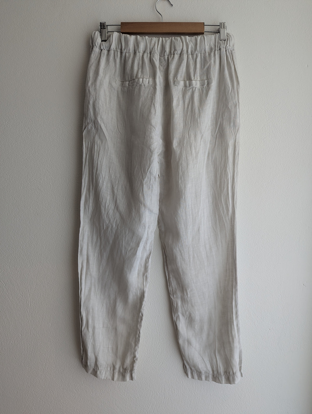 Light beige linen trousers hanging on a wooden hanger against a plain off-white wall.